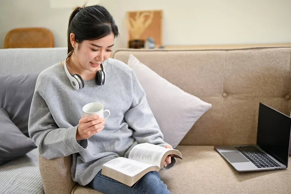迷人而沉稳的千年亚洲女性 一边看书 一边喝咖啡 一边在自家客厅里放松一下 — 图库照片