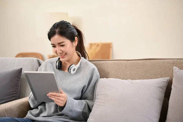 迷人而快乐的千年亚洲女性一边在简约的客厅里放松 一边用数码平板电脑 — 图库照片