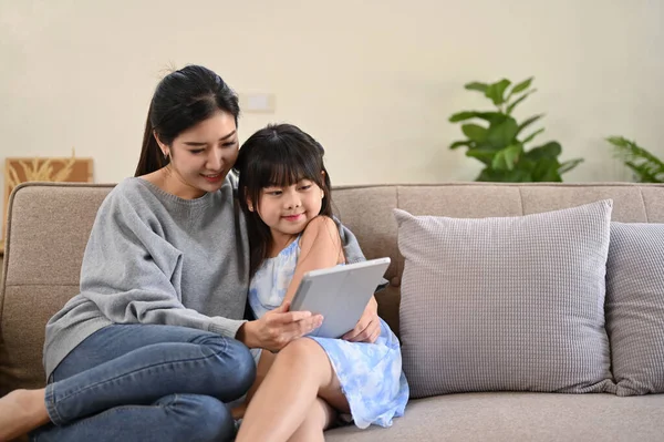 Happy family time concept, A pleased and kind Asian mom using tablet, telling a story, reading an online fairy tale story to her cute daughter while relaxing together in the living room.