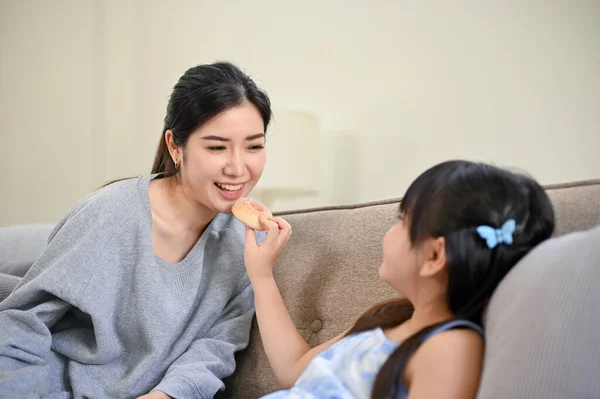 一个漂亮又顽皮的年轻亚洲女孩一边和妈妈一起在客厅里放松 一边喂着甜甜圈 快乐的家庭观念 — 图库照片