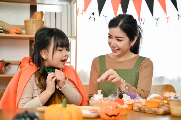 快乐而惊讶的年轻亚洲女孩喜欢在厨房和妈妈一起装饰和做万圣节纸杯蛋糕 — 图库照片