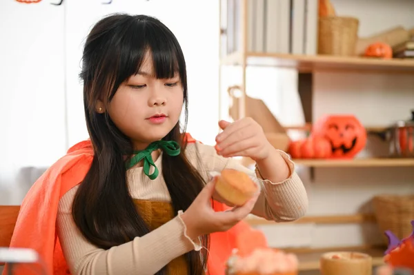 穿着万圣节服装的可爱的亚洲小女孩拿着一个杯子蛋糕 在厨房里装饰着她的杯子蛋糕 — 图库照片