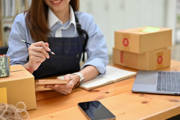 stock image Charming Asian female small online business owner or worker lists her schedule plan on her notepad while preparing shipping orders. cropped shot