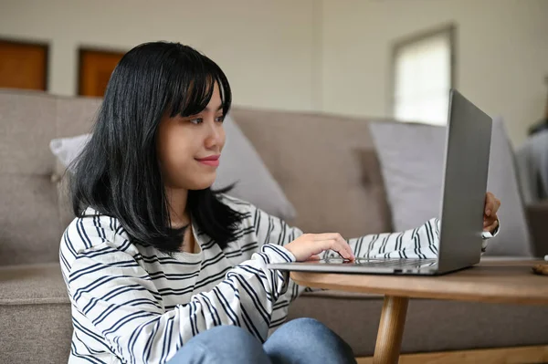 Beautiful young Asian female using her laptop computer in her home living room. work from home concept
