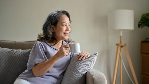 기분좋고 동양인 여성은 거실에서 편안하게 쉬면서 커피를 마시며 표정으로 창밖을 — 스톡 사진