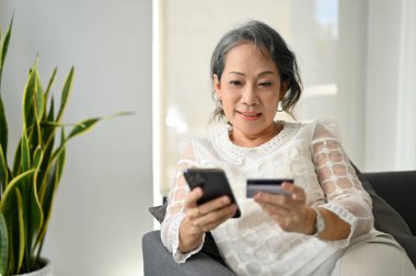 Akıllı telefonu ve kredi kartını kullanarak internet faturalarını ödeyen çekici Asyalı kadın, evden online alışveriş yapmaktan hoşlanıyor..