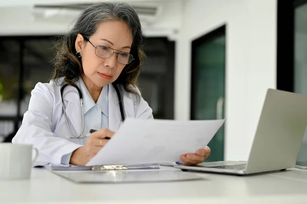 亚洲老年专业女医生 专注于阅读论文上的一个医学研究案例 在她办公室的办公桌前工作 — 图库照片