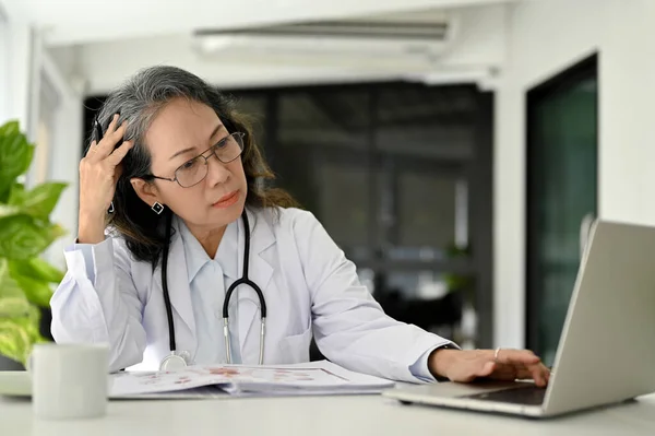 专业的亚洲老年女医生在她的办公桌前工作 在她办公室的笔记本电脑上研究和阅读医学在线研究论文 — 图库照片
