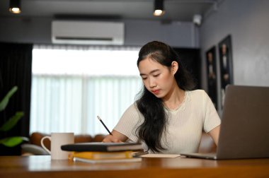 Yetenekli ve güzel Asyalı genç kız üniversite öğrencisi ödevlerini yapmaya, bir kitap hakkında araştırma yapmaya ve not defterine yazmaya odaklanıyor..