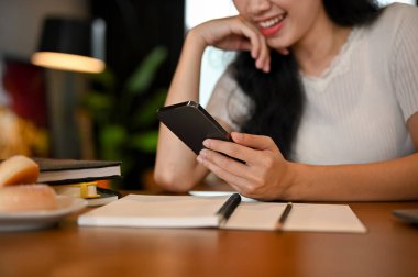 Kesilmiş görüntü, çalışma masasındaki güzel Asyalı genç bayan akıllı telefonunu arkadaşlarıyla sohbet etmek ya da sosyal medya platformunda kaydırmak için kullanıyor..