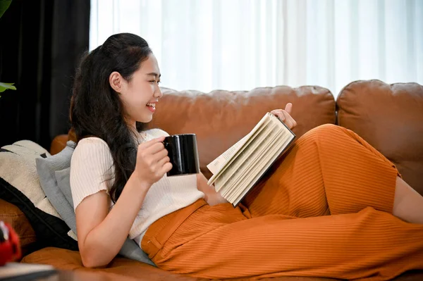 迷人的年轻亚洲女性在沙发上放松一下 一边喝着早晨的咖啡 一边看书或小说 — 图库照片