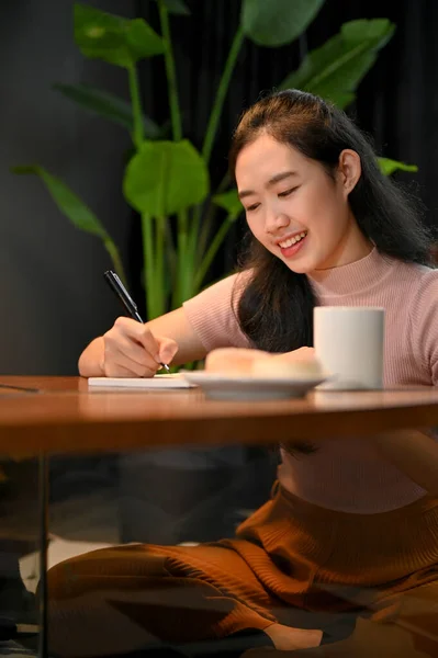 漂亮的年轻亚洲女大学生做功课 在笔记本上写东西 坐在咖啡店里 — 图库照片