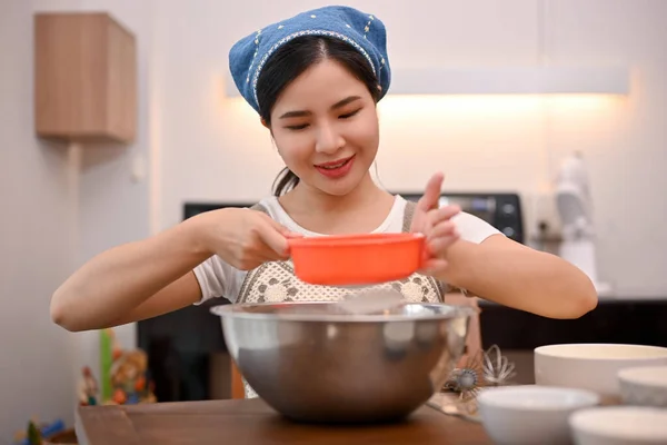 Attractive young Asian female enjoys baking cupcakes in her kitchen, using a flour sifter to prepare the flour in the mixing bowl.