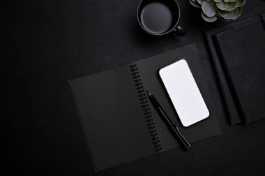 Modern moda siyah çalışma alanı üst görünümü ile akıllı telefon beyaz ekran modeli, kalem, defter, kahve fincanı ve siyah arka planda dekor bitkisi.