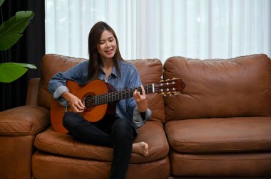 Çekici genç Asyalı kadın klasik gitarını oturma odasında, eski kahverengi deri kanepesinde çalıyor. Hobi ve eğlence konsepti