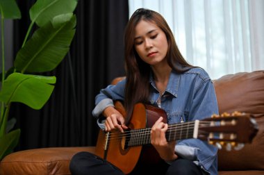 Hipster ve çekici Asyalı kadın kot ceketli modern klasik oturma odasında gitar çalıyor..