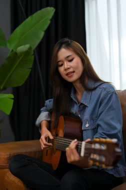 Güzel ve yetenekli kot ceketli Asyalı kız modern oturma odasında akustik gitarını çalarken şarkı söylüyor..