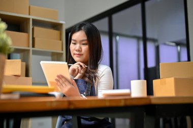 Çekici ve yetenekli Asyalı genç bayan çevrimiçi dükkan sahibi stoklarını kontrol ediyor ve dijital tablet sitesindeki ürün durumunu güncelliyor. KME iş kurma kavramı