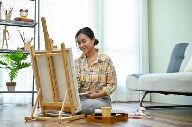 Çekici ve yetenekli genç Asyalı kadın oturma odasında oturuyor, resim yapıyor, tuval sehpasında akrilik renkle resim yapıyor..