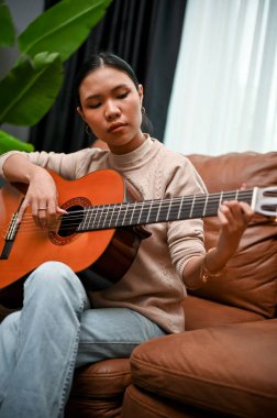 Portre, yetenekli ve kendinden emin bin yıllık Asyalı kadın gitar çalarak rahatlıyor, evde gitar çalma becerisini geliştiriyor..