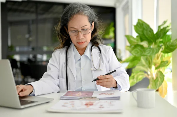亚洲老年专业女医生 专心致志于医学研究 阅读医疗论文 在医生办公室的办公桌前工作 — 图库照片