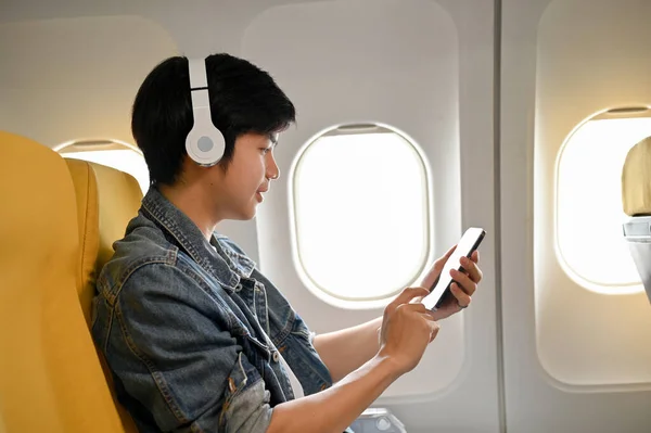 Handsome young Asian male traveler or passenger at the window seat in economy class is using his mobile phone during the flight.