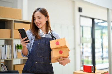 Enfes genç Asyalı bayan mağaza sahibi ya da ticari girişimi akıllı telefonunu kullanarak nakliye paketlerini stok ofisinde tutuyor. E-ticaret girişimi konsepti