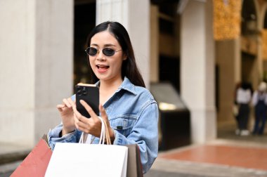Yakışıklı genç Asyalı alışveriş torbaları taşıyan, akıllı telefonunu kullanan, alışveriş merkezinde alışveriş yaparken arkadaşlarıyla sohbet eden bir bayan. şehir yaşam tarzı kavramı