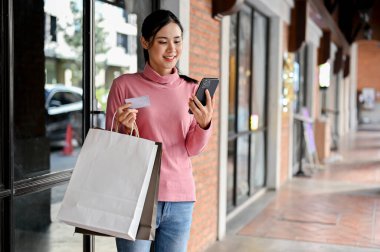 Güzel bir Asyalı kadın kredi kartı tutuyor, akıllı telefon kullanıyor, alışveriş torbaları taşıyor, alışveriş merkezinden alışveriş yapmayı seviyor..