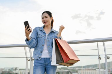 Alışveriş torbaları taşırken akıllı telefon kullanan Asyalı kadın alışveriş merkezinde alışveriş yapmayı seviyor. modern yaşam tarzı kavramı