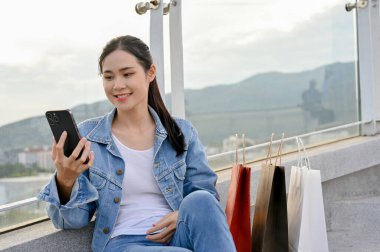 Alışveriş torbalarıyla otururken akıllı telefon kullanan Asyalı kadın, alışveriş merkezinden alışveriş yapmayı seviyor..