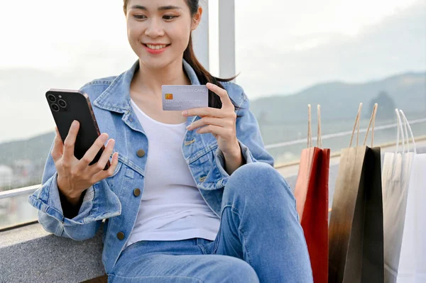 漂亮而快乐的年轻亚洲女人拿着信用卡 拿着购物袋坐在城市购物中心的屋顶上 城市生活方式概念 — 图库照片