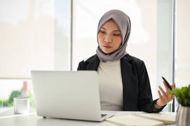 Başarılı Asyalı Müslüman iş kadını tesettür takıyor, dizüstü bilgisayar kullanıyor, görevlerine konsantre oluyor, ofiste çalışıyor..