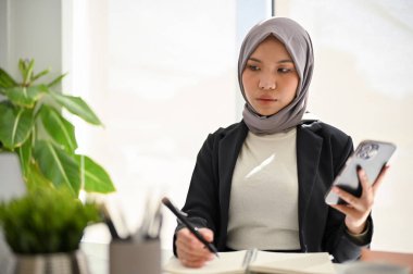 Profesyonel Asyalı Müslüman iş kadını tesettür takıyor, akıllı telefonunu tutuyor, not defterine not alıyor, görevlerine konsantre oluyor..
