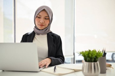 Başarılı ve kendinden emin Asyalı Müslüman iş kadını tesettür takıyor, dizüstü bilgisayar kullanıyor, görevlerine konsantre oluyor, ofiste çalışıyor..