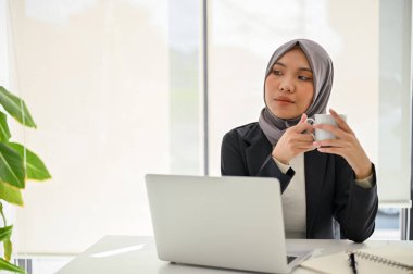 Kendinden emin ve başarılı Asyalı Müslüman iş kadını sabah kahvesini yudumluyor, başarıyı hayal ediyor, ofisinde masasında oturuyor..