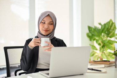 Profesyonel ve muhteşem Asyalı Müslüman iş kadını ya da kadın yönetici bilgisayarın ekranına bakarken kahvesini yudumluyor, özel ofisinde çalışıyor..