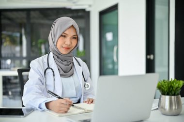 Güzel ve profesyonel, tesettürlü, tıbbi durumuna odaklanan, dizüstü bilgisayar kullanan, tıbbi form dolduran, ofisinde çalışan Müslüman kadın doktor..