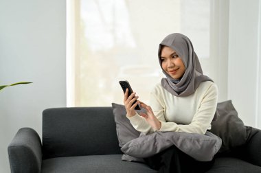 Yakışıklı Asyalı Müslüman kadın, oturma odasındaki kanepede dinlenirken akıllı telefonunu kullanarak tesettür takıyor..