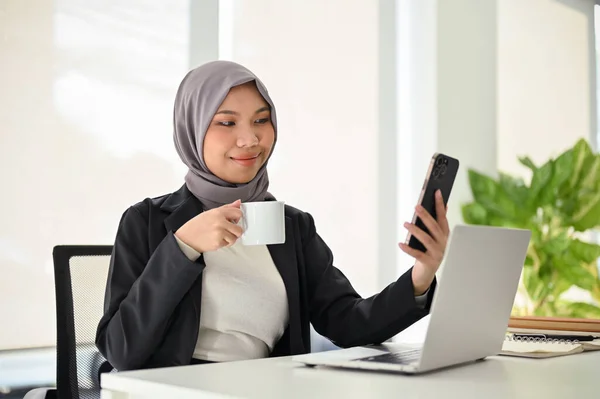Happy and professional Asian Muslim businesswoman or female manager using her smartphone while sipping morning coffee at her desk.