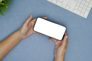 Mavi çalışma alanı arka planında yatay pozisyonda akıllı telefon beyaz ekran modeli tutan bir kadın el. en üstteki görünüm