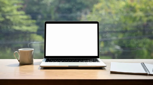 A laptop computer white screen mockup, coffee cup and spiral notepad on the table against the window. coffee shop workplace