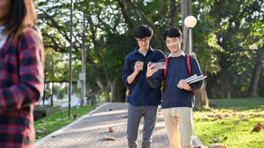 İki genç Asyalı üniversite öğrencisi akıllı telefon ekranına bakıyor, kampüsün park yolunda birlikte yürürken video izliyorlar. en iyi arkadaşlar, yakın arkadaşlar, arkadaşlık