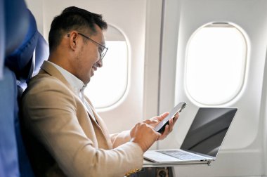 Yan bakış, akıllı ve başarılı Asyalı iş adamı veya CEO uçuş sırasında akıllı telefonunu kullanarak birinci sınıf koltukta oturur..