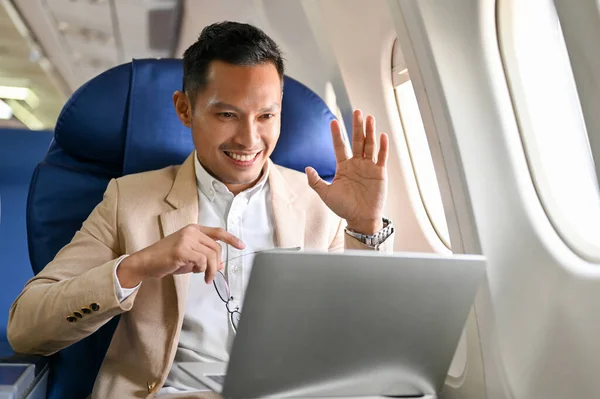 Professional and successful Asian businessman or male CEO waving his hand while having an online meeting or conference with a client during the flight for his overseas business meeting.