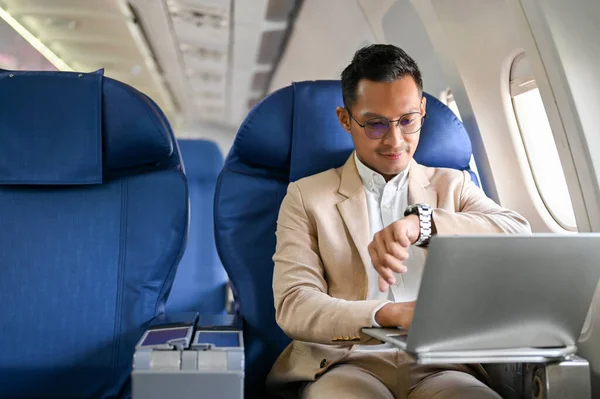 Successful Asian businessman or male CEO sits at the window seat in business class, checking his watch for the arrival time, on the flight for his overseas business trip.