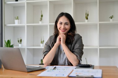 Çekici ve neşeli Asyalı iş kadını masasında oturuyor ellerini birleştiriyor, gülümsüyor ve kameraya bakıyor..