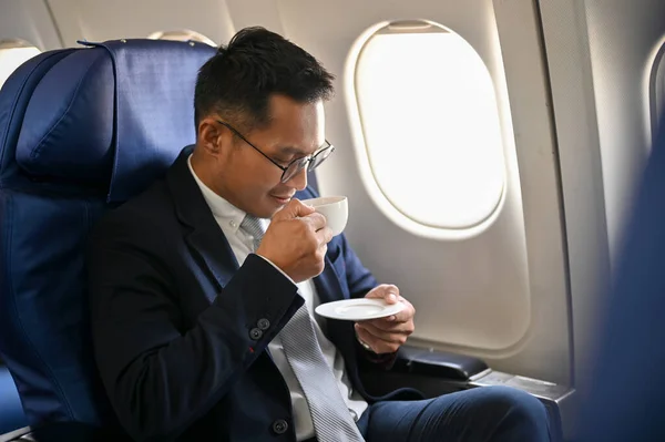 Successful Asian businessman or male CEO sits at the window seat on his private jet sipping coffee during the flight for an overseas business meeting.