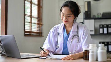 Profesyonel Asyalı doktor ya da beyaz önlüklü eczacı klinikteki masasında görevlerini yapıyor..