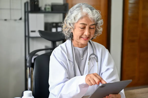 Happy and professional Asian aged female doctor checking her schedule, reviewing medical record on her digital tablet.